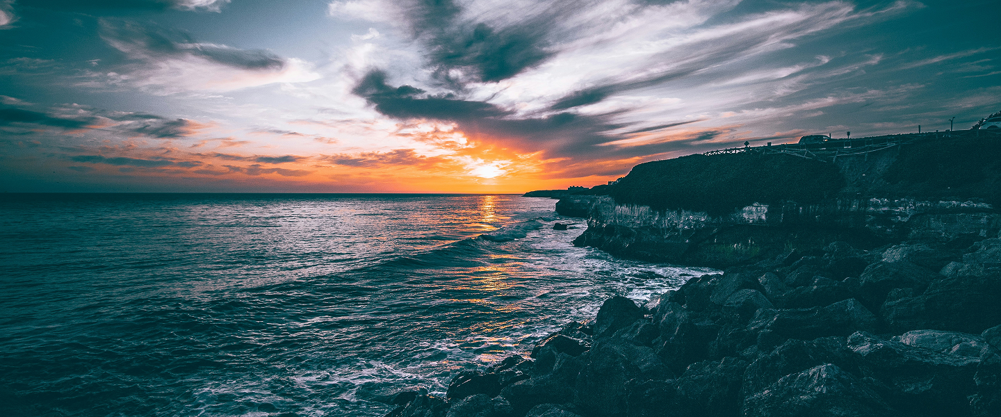 A dramatic sunset over the ocean with vibrant orange and purple hues. Rocky shoreline and dark clouds add an intense, serene atmosphere.